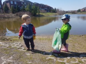 Two toddlers stand beside a body of water. One of them is holding a plastic grocery store bag that is partially full.