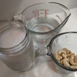 A jar of cashew milk is pictured beside a measuring cup of water and a smaller measuring cup of cashews