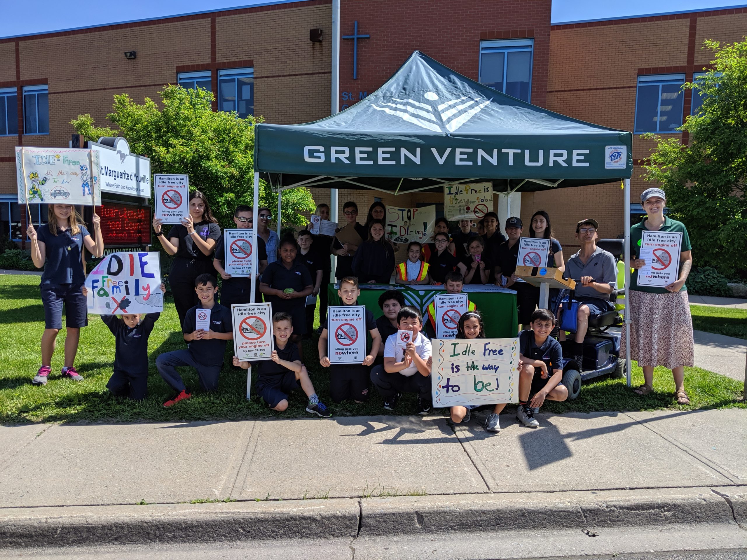 A classroom of children stand and sit in front of a school, holding up no idling signs and posters beneath a Green Venture tent.