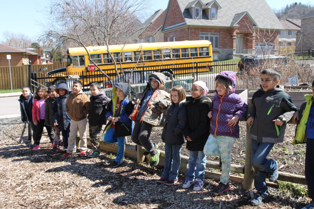 A class of children lean against the EcoHouse community garden fence. A schoolbus sits on the road in the background.
