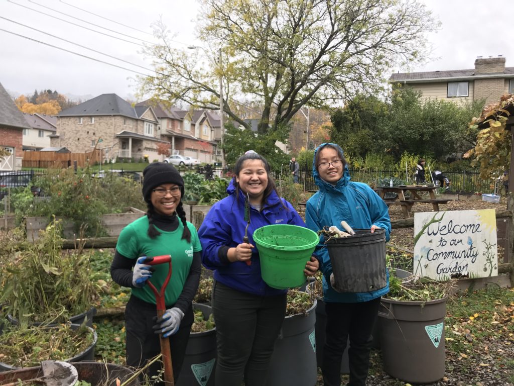 Three youth stand holding a shovel and garden clean-up buckets in the EcoHouse community garden.
