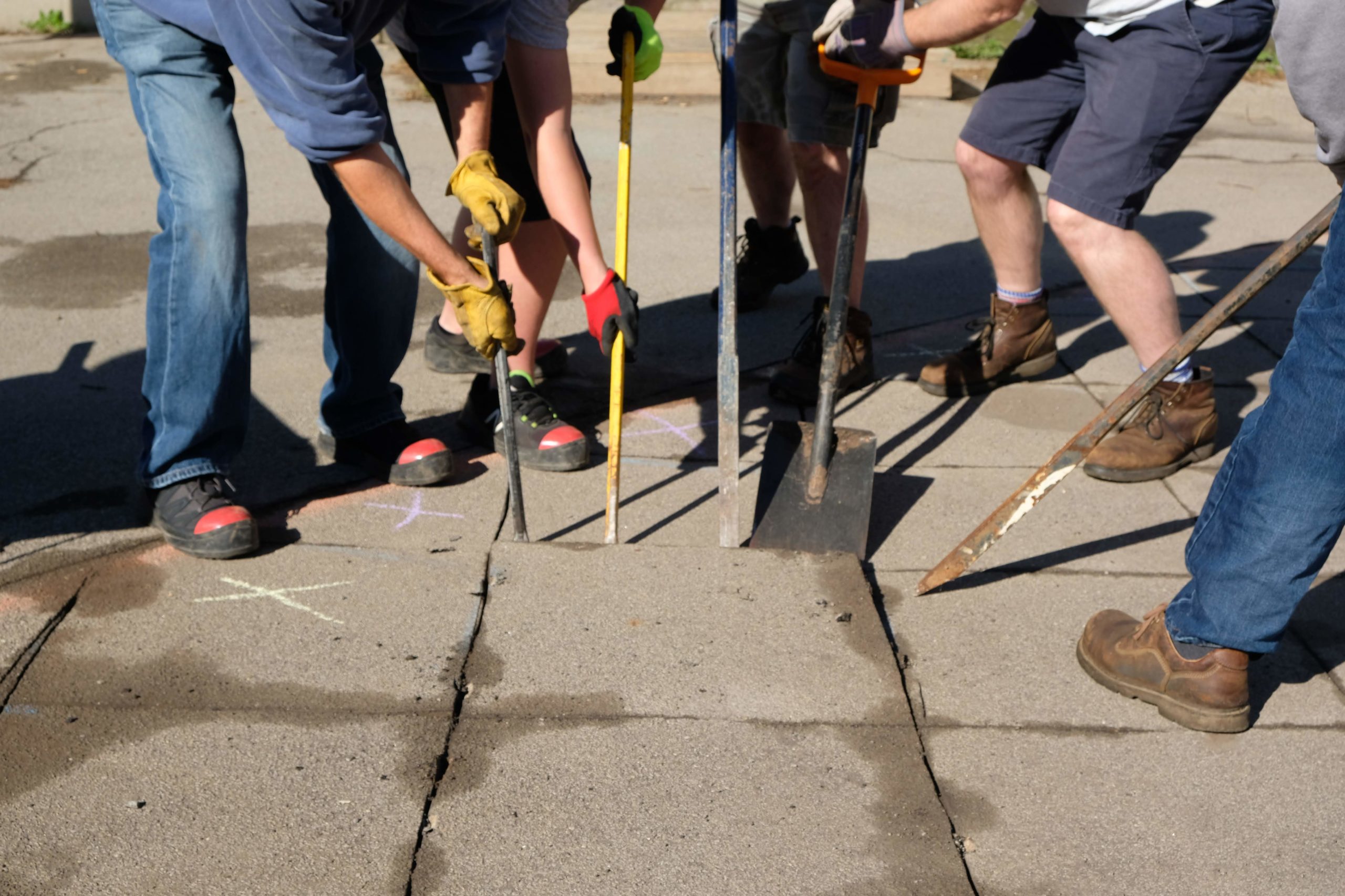 The legs and arms of five people holding shovels and tools work to lift a concrete paver out of the ground.