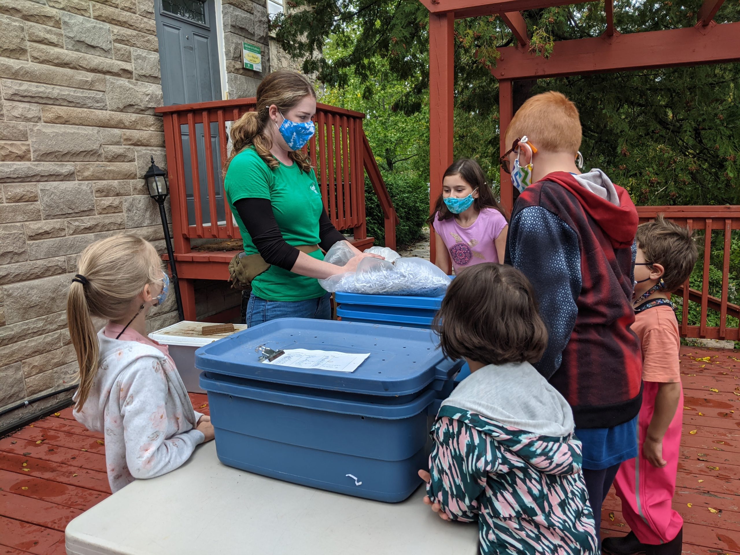 Picture of a masked Green Venture Staff Member who is standing outdoors in front of two blue storage bins that have been turned into vermicomposters. She is surrounded by a group of masked young children.