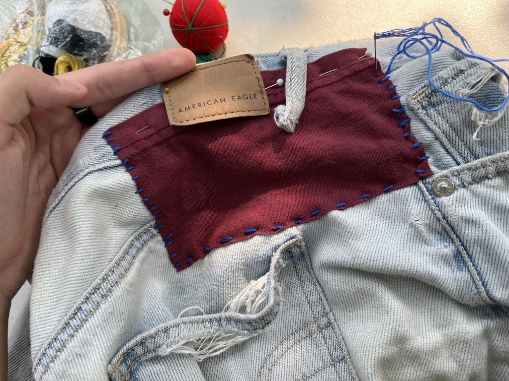 [ID: Red fabric patch on denim shorts. It is partially sewn on with blue thread. The rest of the patch is held down with pins.]