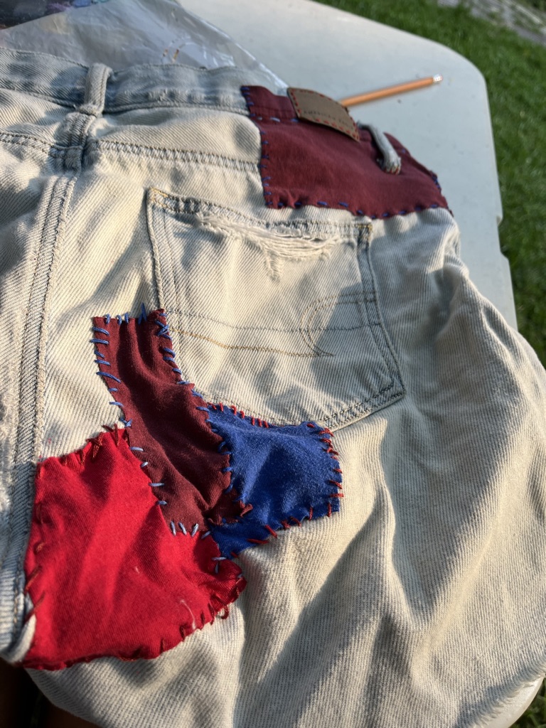 [ID: Denim shorts with four patches sewn on. The patches are and thread are shades of red and blue]