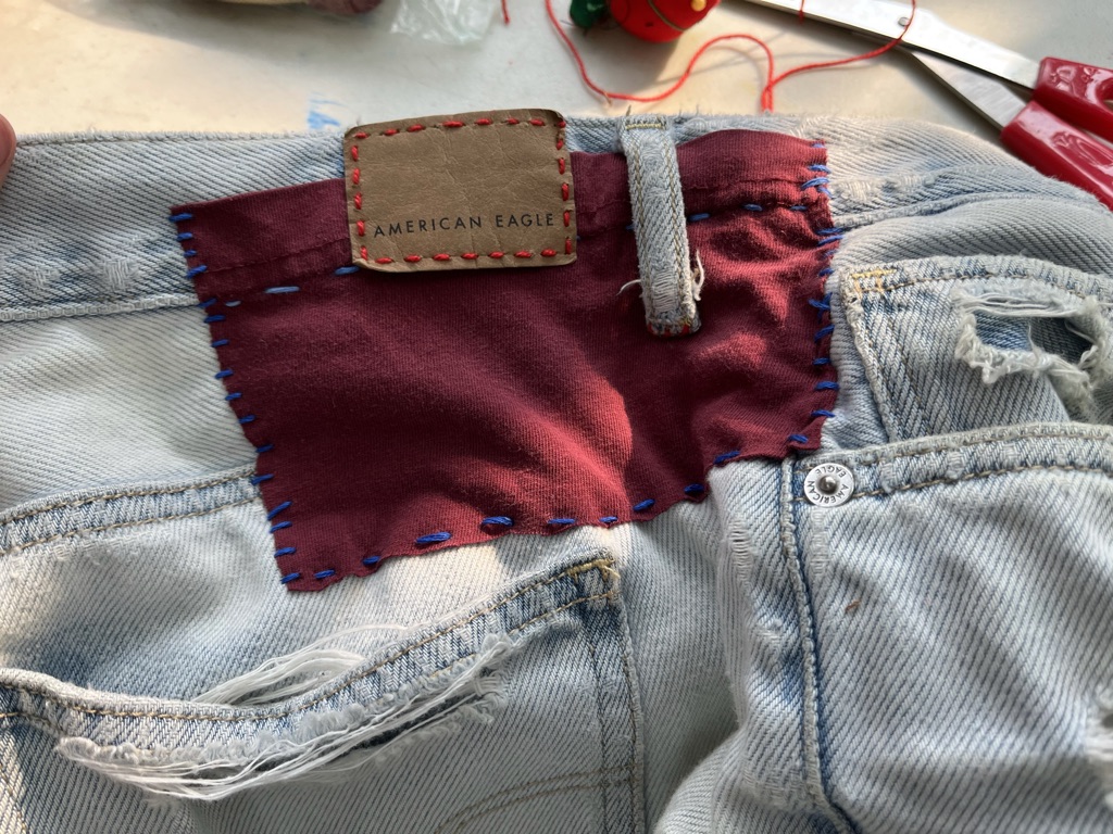 [ID: Denim shorts with red patch sewn on with blue thread and leather label sewn on with red thread]