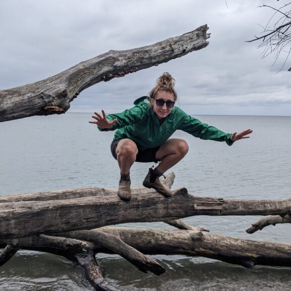 Sarah squats on a fallen tree by a lake on a cloudy day