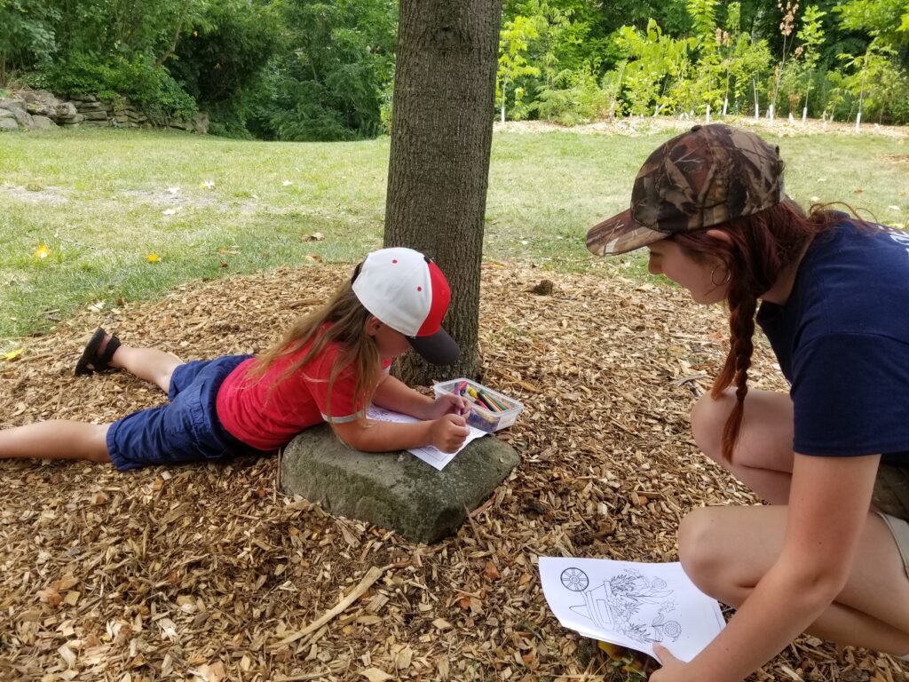 A young child coloring outside under a tree with a leader