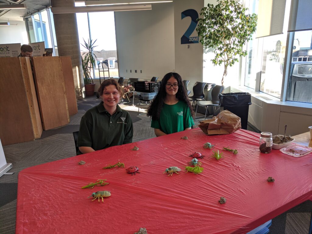 Volunteers Jasmine and Jordyn sit at a table with toy insects